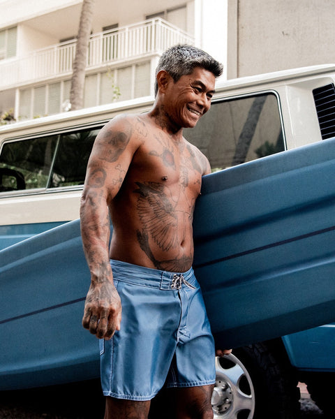 A smiling, tattooed man with gray hair stands shirtless in 310 Boardshorts - Federal Blue, holding a blue surfboard by a van. An apartment building and palm tree are visible in the background.