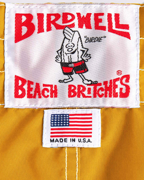 A white patch with red text reads “Birdwell Beach Britches” above a cartoon bird in red shorts, with a U.S. flag patch and “Made in U.S.A.” on durable gold SurfNyl fabric—classic Birdwell 310 Boardshorts.