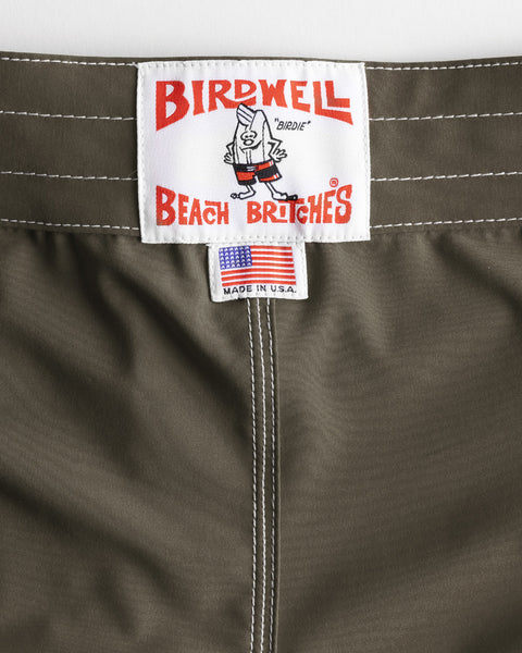 Close-up of the 806 SurfStretch Side Pocket Boardshorts - Army waistband with white stitching, showcasing a Birdwell Beach Britches label with a cartoon bird and a small tag featuring an American flag and “Made in U.S.A.” text.