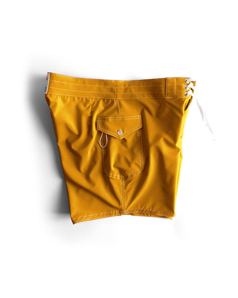 The 806 SurfStretch Side Pocket Boardshorts in gold feature white stitching, a side pocket with button flap and key loop, a white lace-up tie, and are made in the USA. Shown against a white background.