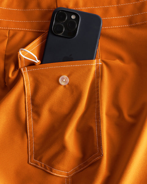 A black smartphone is partially tucked into the buttoned back pocket of 806 SurfStretch Side Pocket Boardshorts in medium orange, featuring white stitching and made in the USA.