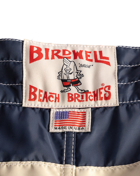 Close-up of a Birdwell Beach Britches label with a bird mascot in red shorts. Below, a smaller MADE IN U.S.A. tag with an American flag highlights the 300 Comp Stripe - Navy/Bone SurfNyl boardshorts in blue and cream.