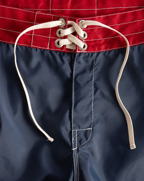 Close-up of the 300 Legend - Navy/Red/Bone boardshorts waistband, made in USA, showing red fabric on top, navy blue below, and a white lace-up drawstring threaded through metal eyelets.