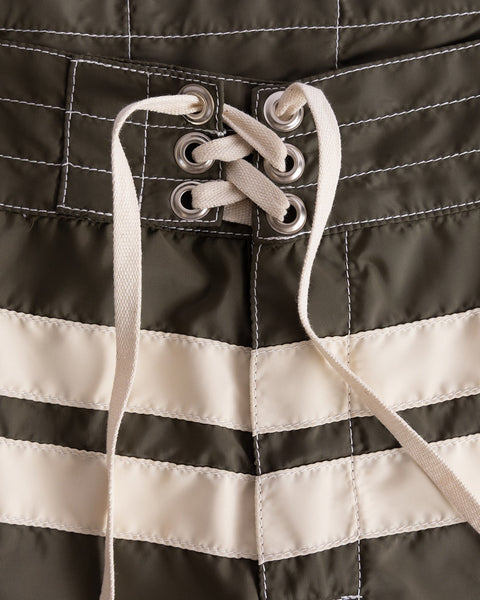 Close-up of 300 Comp Stripe boardshorts in Dark Olive/Bone, featuring white stitched lines, horizontal stripes, and a white lace-up drawstring threaded through metal eyelets at the waist.
