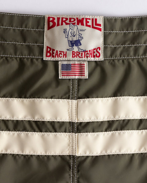 Close-up of the 300 Comp Stripe - Dark Olive/Bone boardshorts, featuring two white stripes, an American flag patch, and the Birdwell Beach Britches label—crafted from SurfNyl and proudly made in the USA.