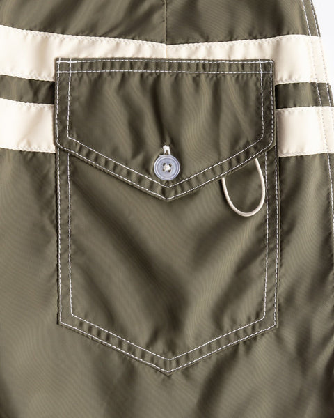 Close-up of the 300 Comp Stripe - Dark Olive/Bone boardshorts pocket with white stitching, flap with button, two beige stripes above, silver D-ring below the flap; proudly made in the USA.