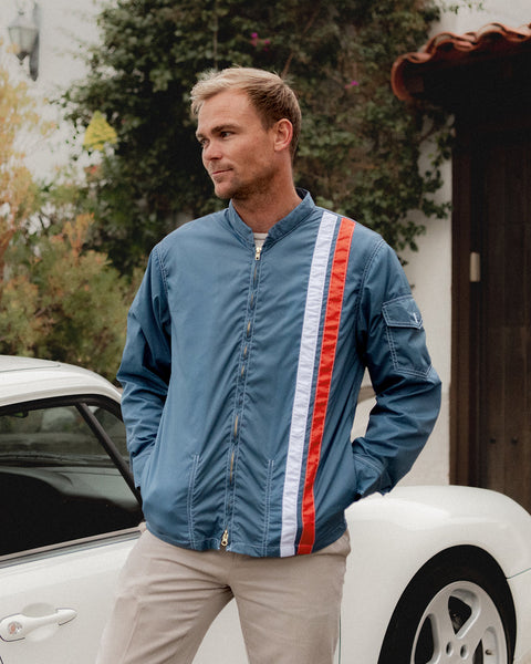 Christian Wach wearing the Racing Jacket 25 Le Mans in Federal Blue with two vertical racing stripes standing in front of a white Porche with white home in background with Green ivy and greenery on wall and wooden brown door with red ceramic shingles.